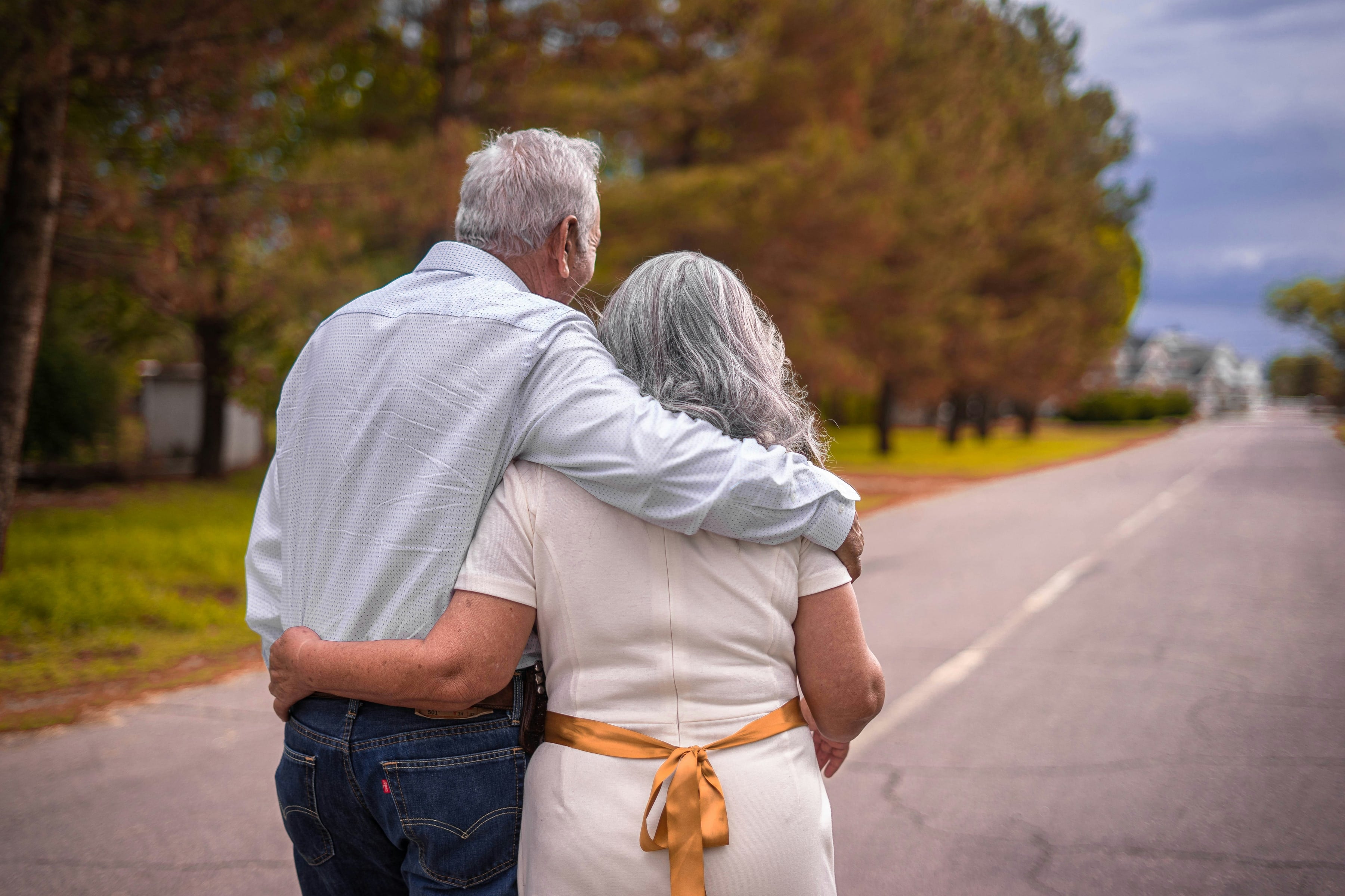 An older couple, seen from behind, walking arm in arm down a quiet tree-lined road