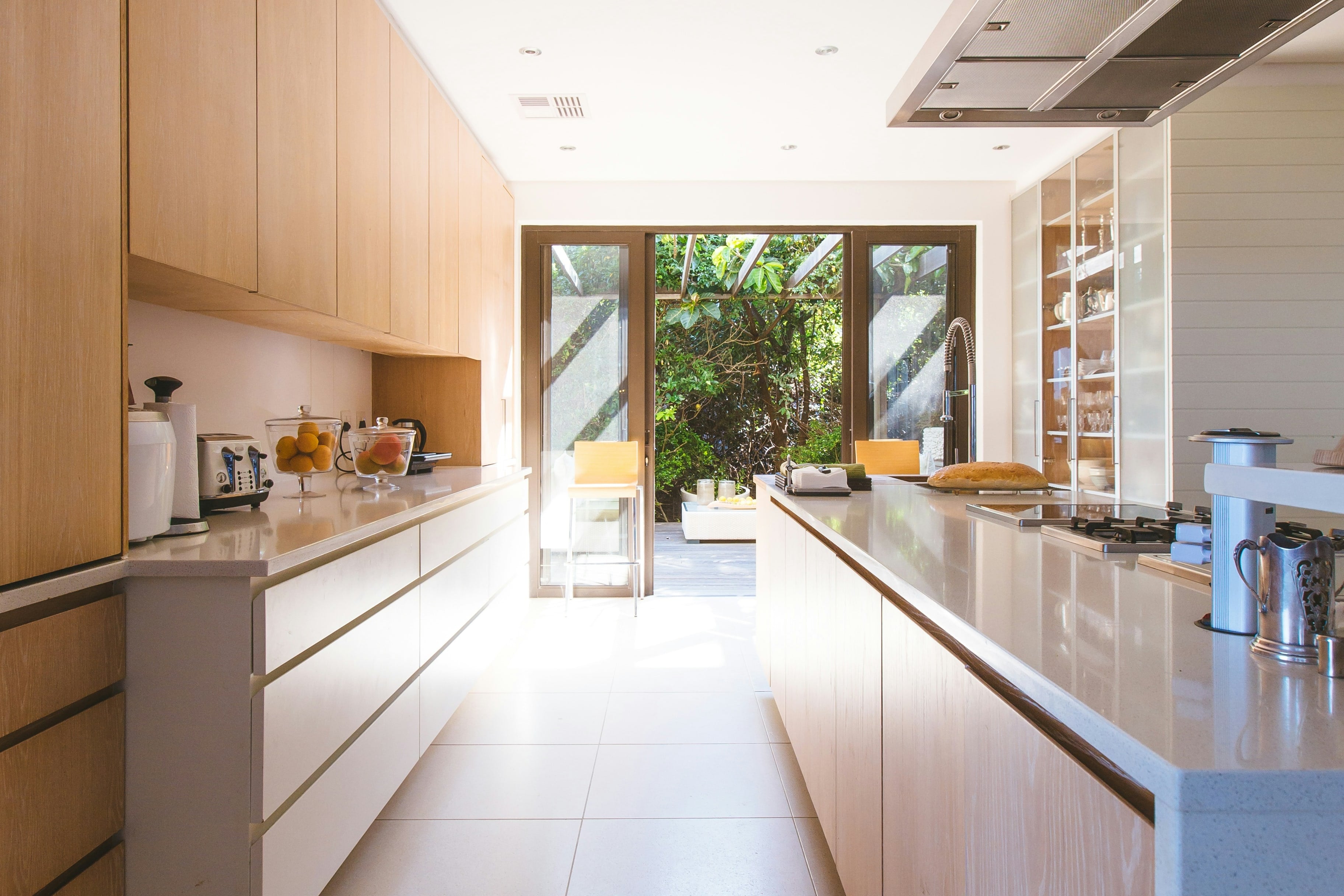 A sunlit kitchen with warm wood cabinets, looking out through open glass doors to a green garden